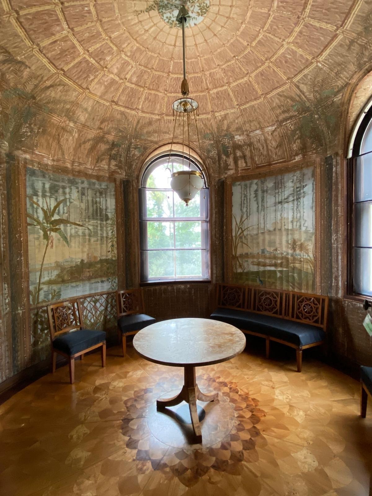 Ein Zimmer im Schloss. Die Wände sind mit Bildern der umgebenden Natur bemalt, der Boden ist aus Holz und mit Intarsien, ein runder Tisch und Stühle sind vor einem Fenster angeordnet.